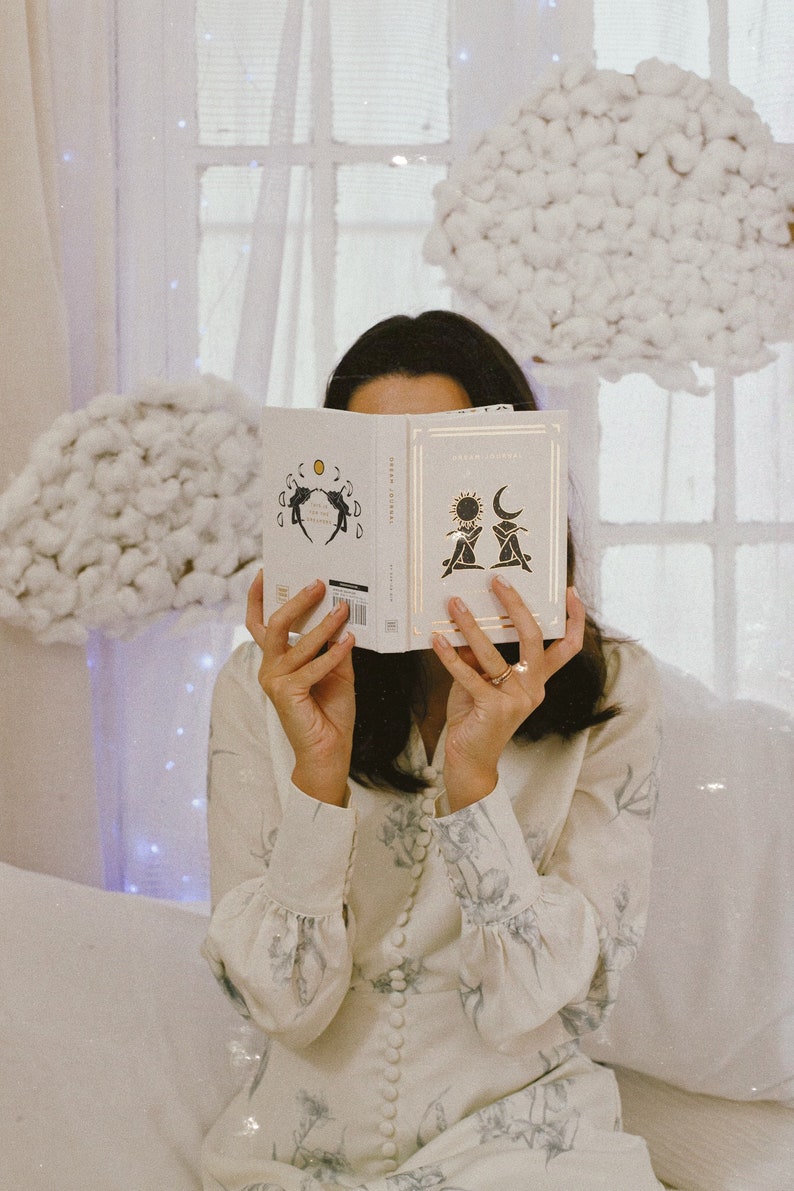 May include: A person is holding a white book open with a black and white illustration of two figures with a sun and moon above them. The book's title is "Dream Journal".