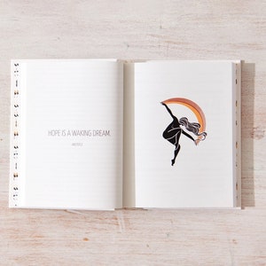 May include: An open journal with a quote on the left page that reads "Hope is a waking dream." The right page features a black silhouette of a person dancing with a rainbow arc behind them.