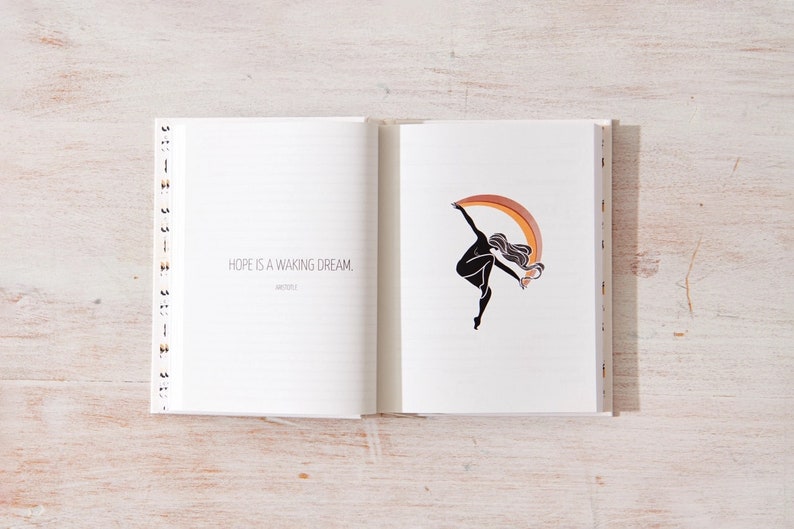 May include: An open journal with a quote on the left page that reads "Hope is a waking dream." The right page features a black silhouette of a person dancing with a rainbow arc behind them.