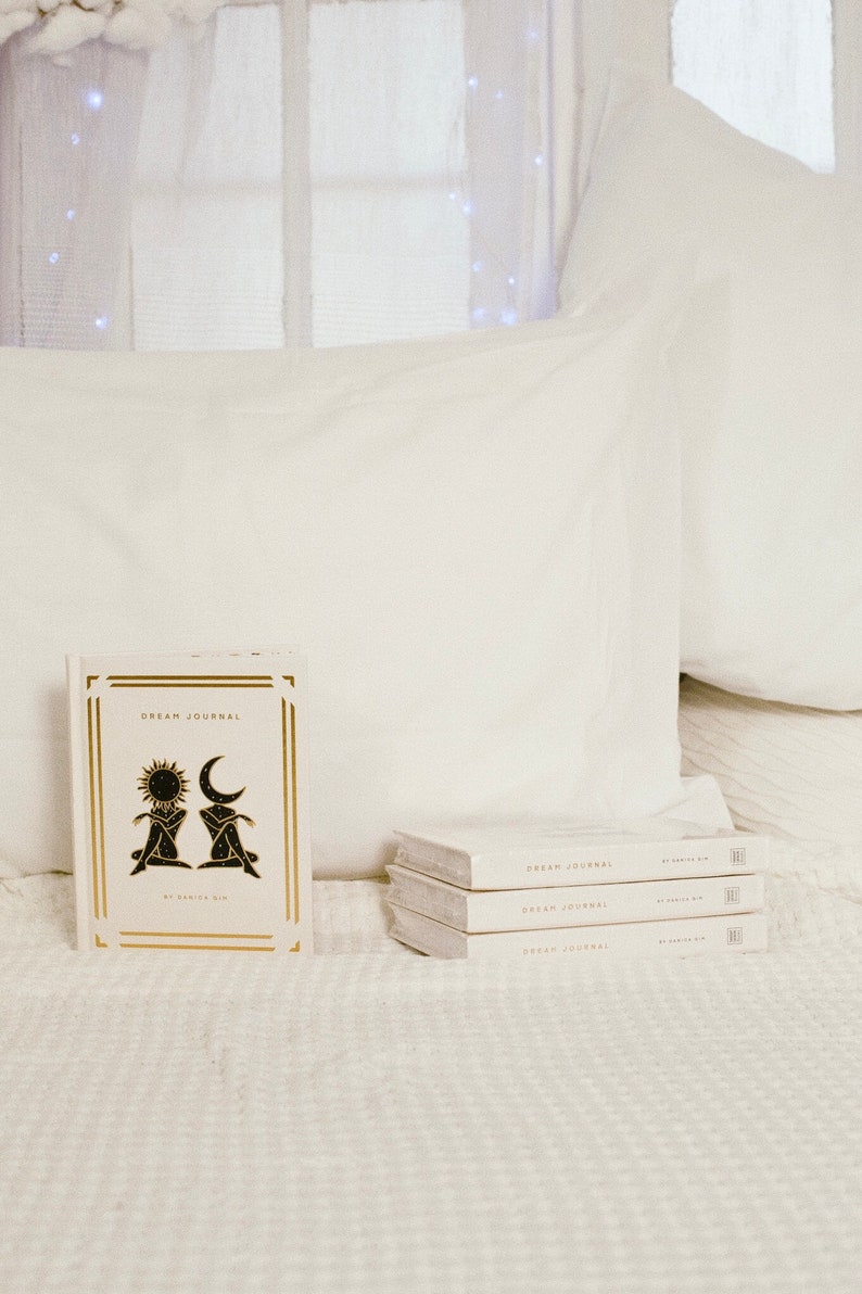 May include: A stack of three white dream journals with gold lettering that reads "Dream Journal" on the front cover. The top journal has a gold illustration of two figures with a sun and moon above them.