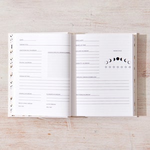 May include: A white journal with a black and white moon phase design on the cover. The journal is open to a page with a section for recording dreams. The page includes sections for date, dream title, emotions felt in the dream, people in the dream, dream setting, animals in the dream, flowers in the dream, recurring dream elements, specific dream symbols, patterns in the dream, and a moon phase chart.