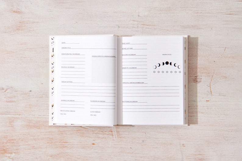 May include: A white journal with a black and white moon phase design on the cover. The journal is open to a page with a section for recording dreams. The page includes sections for date, dream title, emotions felt in the dream, people in the dream, dream setting, animals in the dream, flowers in the dream, recurring dream elements, specific dream symbols, patterns in the dream, and a moon phase chart.