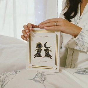 May include: A white dream journal with gold accents and a black illustration of a sun and moon. The text on the cover reads "Dream Journal" and "By Danica Gin".