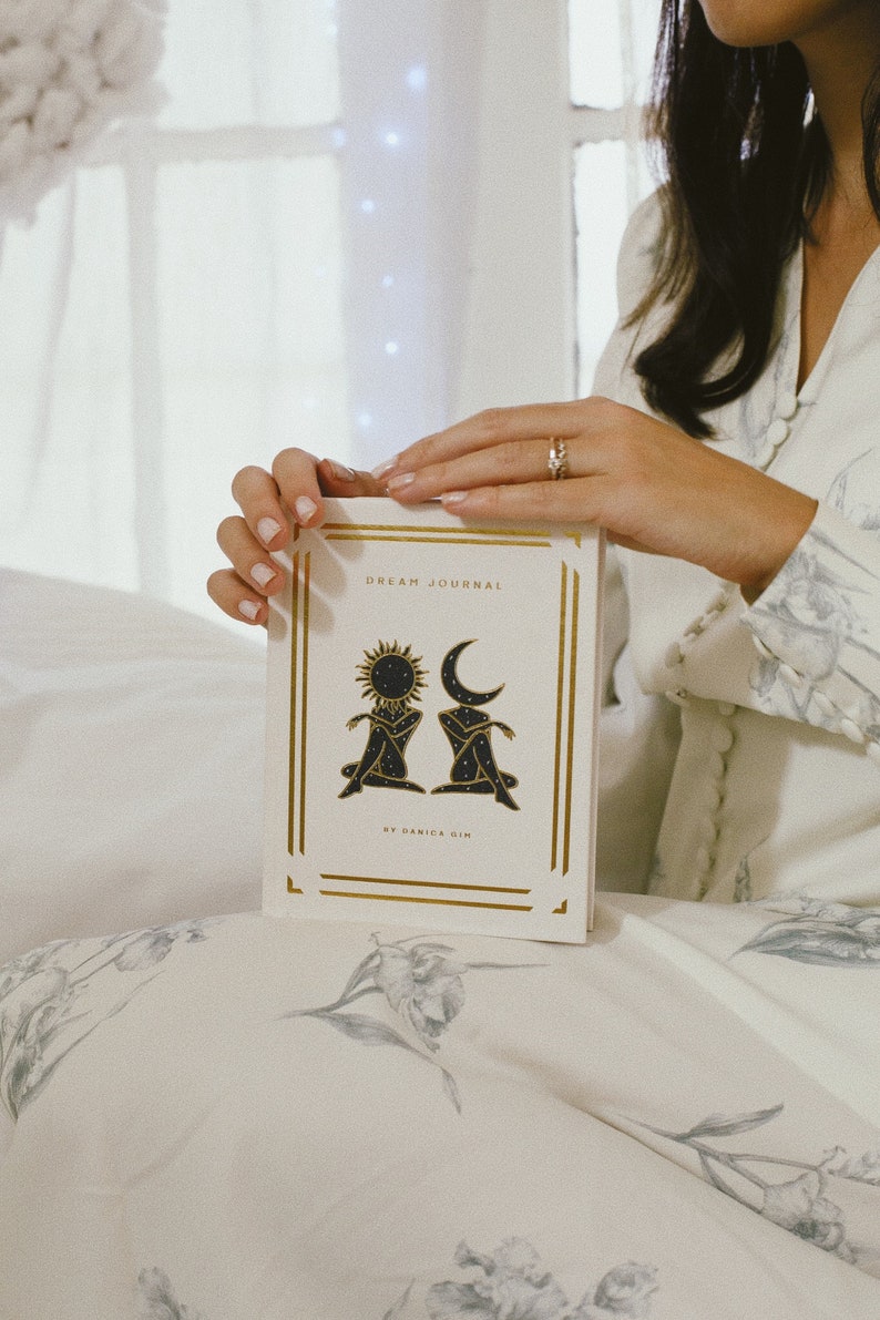 May include: A white dream journal with gold accents and a black illustration of a sun and moon. The text on the cover reads "Dream Journal" and "By Danica Gin".