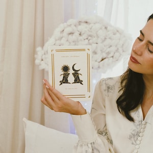 May include: A white journal with gold lettering that reads "DREAM JOURNAL" and a black and gold illustration of a sun and moon. The journal is being held by a person wearing a white floral dress.