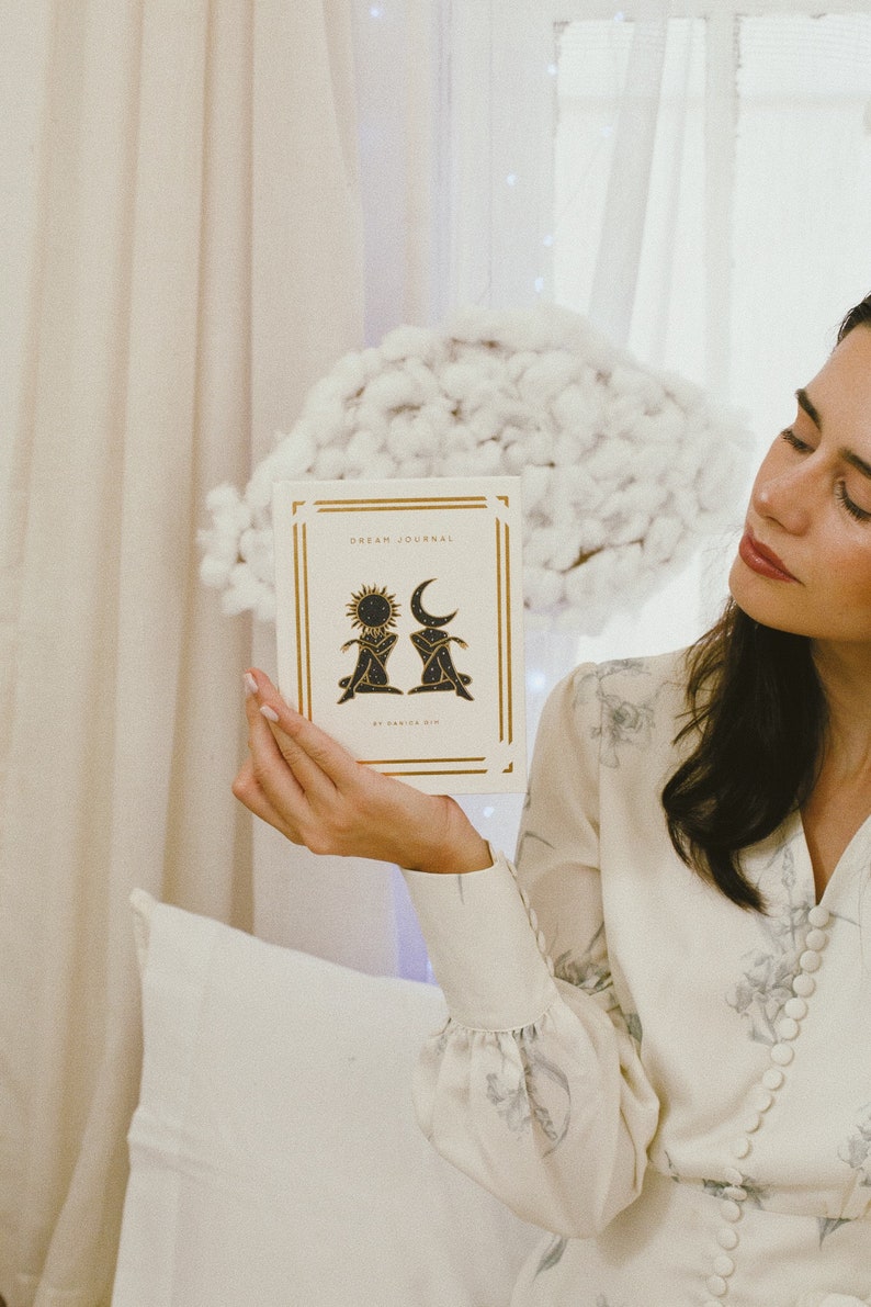 May include: A white journal with gold lettering that reads "DREAM JOURNAL" and a black and gold illustration of a sun and moon. The journal is being held by a person wearing a white floral dress.