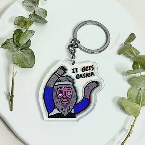 Könnte beinhalten: Ein silberner Schlüsselanhänger mit einem transparenten Acryl-Anhänger, der eine Cartoon-Illustration einer Person mit Bart und dem Text "IT GETS EASIER" zeigt.