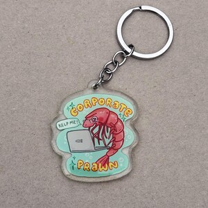 May include: A clear acrylic keychain featuring a cartoon prawn working on a laptop. The prawn is red with glasses, and the keychain has the words "CORPORATE PRAWN" and "KELP ME!" in a speech bubble. A silver keyring is attached.