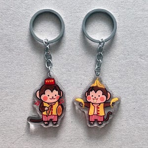 May include: Two monkey-themed keychains. One keychain features a monkey playing a drum and cymbals, and the other holds bananas. Both monkeys wear red and yellow outfits and have silver key rings.