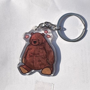 Peut inclure: Un porte-clés en acrylique transparent avec une illustration d'ours brun et des accents de fleurs roses. L'ours a un corps rond et est assis. Le porte-clés est doté d'un anneau en métal argenté pour l'attacher.