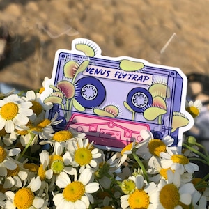 May include: A purple and pink cassette tape sticker with the text "Venus Flytrap" on it. The tape is surrounded by illustrations of Venus flytraps.