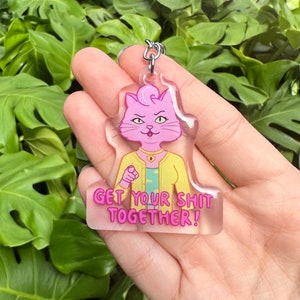 Könnte beinhalten: Ein klarer Acryl-Schlüsselanhänger mit einer rosa Katzenfigur, die eine gelbe Jacke trägt, und dem rosa Text "GET YOUR SHIT TOGETHER!". Der Schlüsselanhänger hat einen silbernen Metall-Schlüsselring.