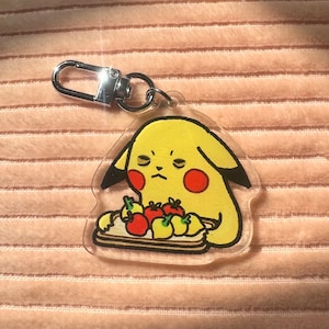 Può includere: Un personaggio dei cartoni animati giallo con le guance rosse che tiene un vassoio di frutta rossa e gialla. Il personaggio tiene il vassoio con entrambe le mani e ha un'espressione scontrosa sul viso. Il personaggio è un portachiavi in ​​acrilico trasparente.
