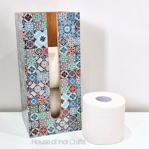 May include: A decorative tissue box with a colorful tile pattern, next to a roll of white toilet paper. The tissue box has a wooden interior and a rectangular shape. The tile design includes blue, green, red, and gray squares.