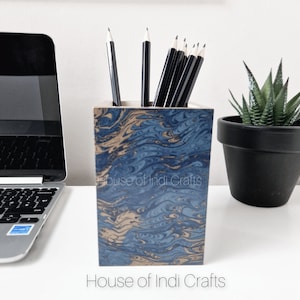 Könnte beinhalten: Ein quadratischer Stifthalter mit blauem und goldenem Marmormuster, gefüllt mit schwarzen Bleistiften. Der Stifthalter trägt den Text "House of Indi Crafts". Ein Laptop und eine Topf-Sukkulente befinden sich im Hintergrund.