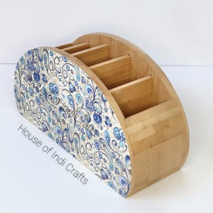 Può includere: Un organizzatore in bambù a forma di semicircolo con cinque scomparti. La parte anteriore è rivestita con un carta a motivi blu e bianchi con il testo "House of Indi Crafts".