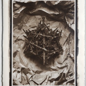May include: A black and white photograph of a pile of rusty nails arranged in a cross shape on a crumpled piece of brown paper.