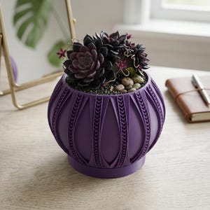 May include: A round, deep purple planter with a textured, ribbed exterior featuring vertical panels and intricate braided detailing. The planter holds a cluster of dark purple succulent plants nestled among small gray pebbles and tiny magenta flowers.