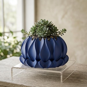 Peut inclure: Un pot de fleurs bleu foncé au fini mat, doté d'un motif ondulé vertical rappelant des pétales. Le pot contient un groupe de plantes succulentes vertes nichées dans de petits cailloux gris foncé, formant un arrangement dense et arrondi.