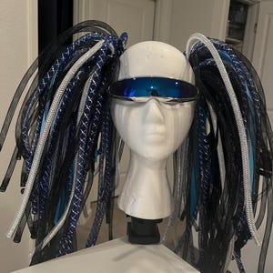 Puede incluir: Una cabeza de maniquí de espuma blanca con gafas de sol azules espejadas y dos coletas grandes. Las coletas están hechas de malla y cinta negras, plateadas y azules. Las gafas de sol son de estilo futurista.