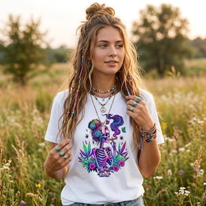 May include: White t-shirt with a vibrant skeleton design smoking a cigarette, encircled by flowers and cannabis leaves. The skeleton has a rainbow-coloured bun and is exhaling purple smoke. The shirt is worn by a person in a field.