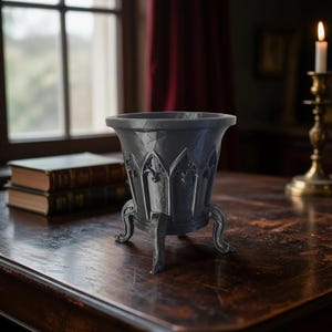 Puede incluir: Una maceta de color gris oscuro, de estilo gótico, con un borde acampanado y tres patas ornamentadas. La maceta presenta detalles arqueados y se exhibe sobre una mesa de madera. Una vela encendida y libros en el fondo.