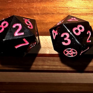 May include: Two black, multi-sided dice with pink numbers and symbols. The dice are set against a wooden background. One die shows the number 2, while the other displays the number 3 and a pentagram symbol.