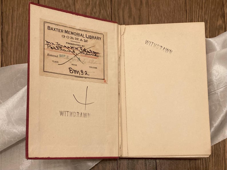 Puede incluir: Un libro abierto con lomo rojo y p&aacute;ginas envejecidas. La p&aacute;gina izquierda presenta una etiqueta de biblioteca con texto, incluyendo "Baxter Memorial Library" y "Withdrawn" estampado debajo. La p&aacute;gina derecha est&aacute; en blanco, tambi&eacute;n estampada con "Withdrawn".