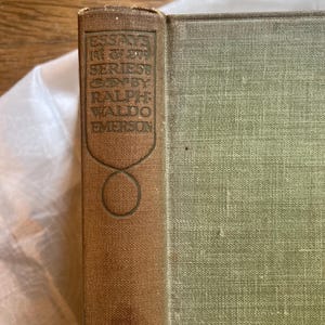May include: A vintage book with a brown spine and green cover. The spine features embossed text that reads "ESSAYS IN FIRST SERIES BY RALPH WALDO EMERSON". The book's cover has a textured appearance.