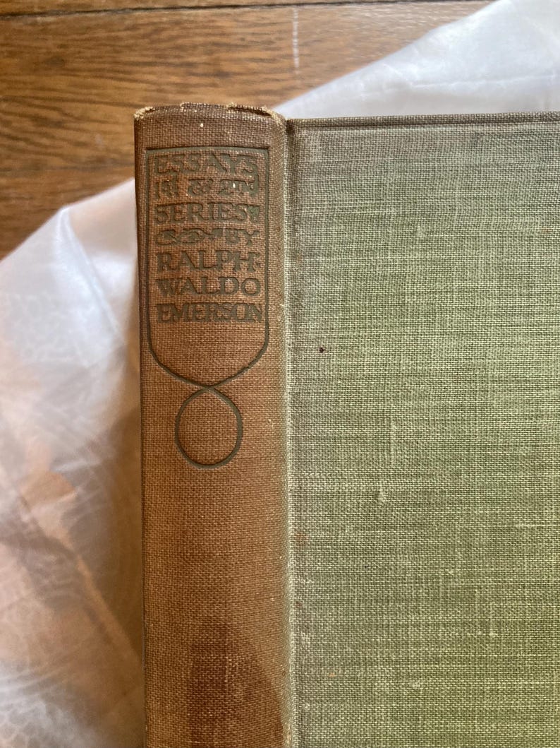 May include: A vintage book with a brown spine and green cover. The spine features embossed text that reads "ESSAYS IN FIRST SERIES BY RALPH WALDO EMERSON". The book's cover has a textured appearance.