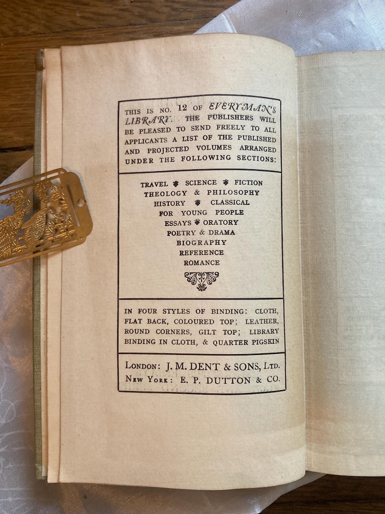 May include: Open book page with text from Everyman's Library, listing sections like Travel, Science Fiction, and History. The text also mentions binding styles such as cloth and leather. A gold-colored bookmark is visible.