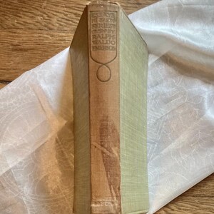 May include: A vintage book with a light green cover and a brown spine. The spine features the title "Essays in First Series" by Ralph Waldo Emerson. The book is resting on a white, patterned cloth.