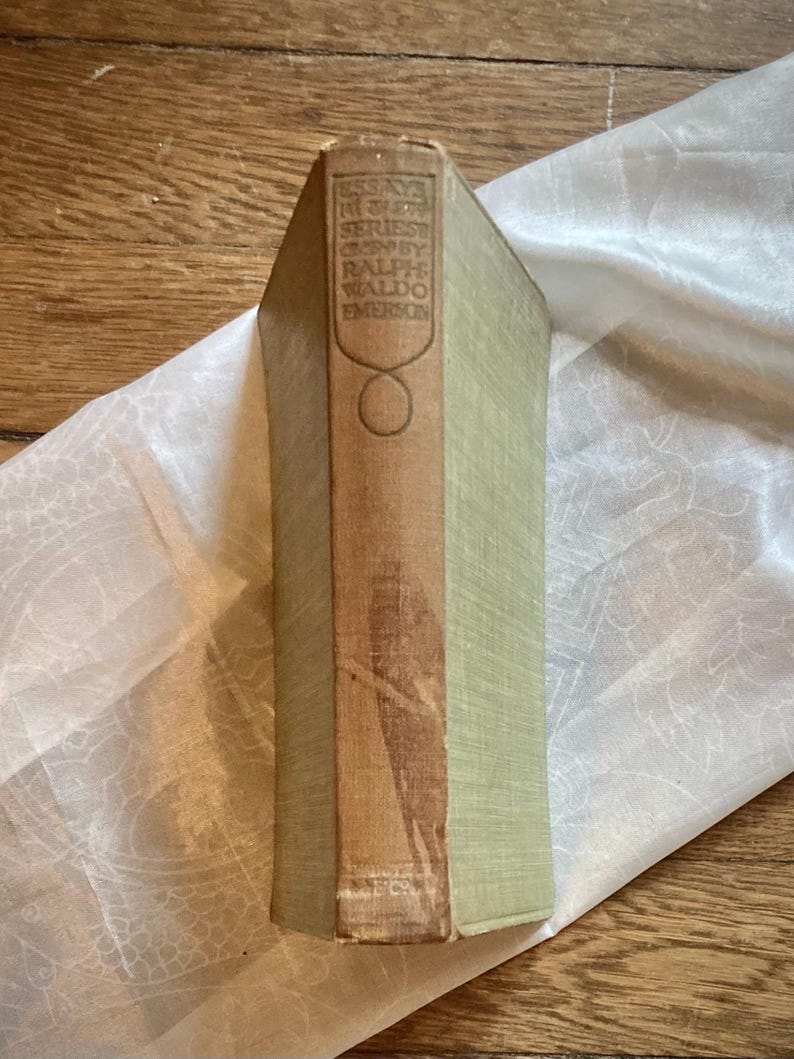 May include: A vintage book with a light green cover and a brown spine. The spine features the title "Essays in First Series" by Ralph Waldo Emerson. The book is resting on a white, patterned cloth.