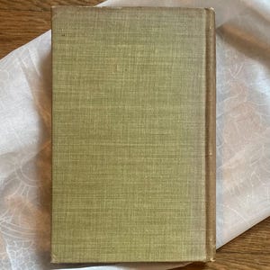 May include: A vintage book with a textured, olive green cover and a brown spine. The book is resting on a white, patterned fabric, with a wooden surface in the background. The book's cover shows signs of wear.