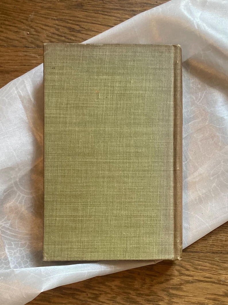 May include: A vintage book with a textured, olive green cover and a brown spine. The book is resting on a white, patterned fabric, with a wooden surface in the background. The book's cover shows signs of wear.