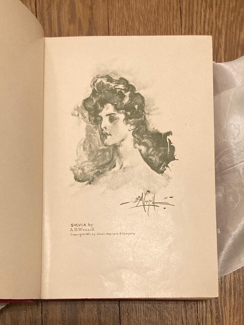 Puede incluir: P&aacute;gina de libro abierta con una ilustraci&oacute;n en blanco y negro de un retrato de mujer. El texto "SYLVIA by A.B. Wenzell" est&aacute; impreso debajo de la imagen, junto con informaci&oacute;n de derechos de autor.