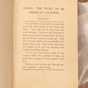Puede incluir: P&aacute;gina de libro abierto con el t&iacute;tulo "SYLVIA: THE STORY OF AN AMERICAN COUNTESS". El cap&iacute;tulo 1 describe un encuentro en Venecia. El texto es negro sobre papel crema. El libro est&aacute; sobre una superficie de madera.