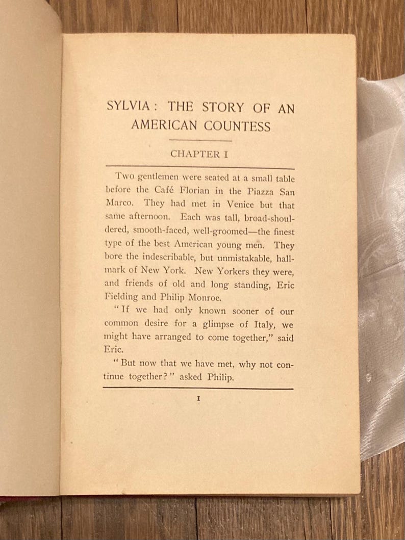 Puede incluir: P&aacute;gina de libro abierto con el t&iacute;tulo "SYLVIA: THE STORY OF AN AMERICAN COUNTESS". El cap&iacute;tulo 1 describe un encuentro en Venecia. El texto es negro sobre papel crema. El libro est&aacute; sobre una superficie de madera.