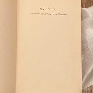 Puede incluir: Libro abierto con el t&iacute;tulo "SYLVIA" y el subt&iacute;tulo "The Story of an American Countess" impresos en negro sobre una p&aacute;gina color crema. El libro est&aacute; abierto en una doble p&aacute;gina, con el texto centrado.