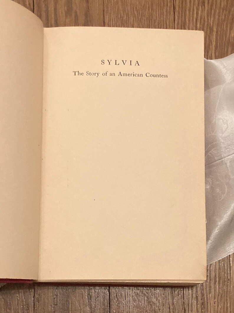 Puede incluir: Libro abierto con el t&iacute;tulo "SYLVIA" y el subt&iacute;tulo "The Story of an American Countess" impresos en negro sobre una p&aacute;gina color crema. El libro est&aacute; abierto en una doble p&aacute;gina, con el texto centrado.