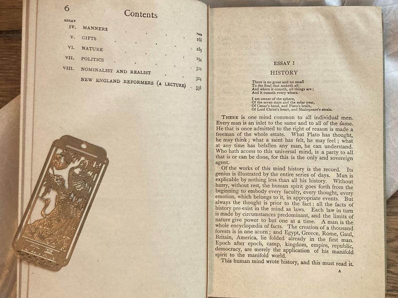 May include: Open book with text and a decorative gold bookmark. The bookmark features an ornate design with figures and a rectangular shape. The book's pages display text from an essay titled "History".