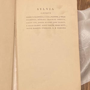 Puede incluir: P&aacute;gina de libro abierta con el t&iacute;tulo "SYLVIA" y una lista de nombres debajo, incluyendo Albert D. Blashfield y Howard Chandler Christy. El texto est&aacute; en una fuente serif en una p&aacute;gina color crema.