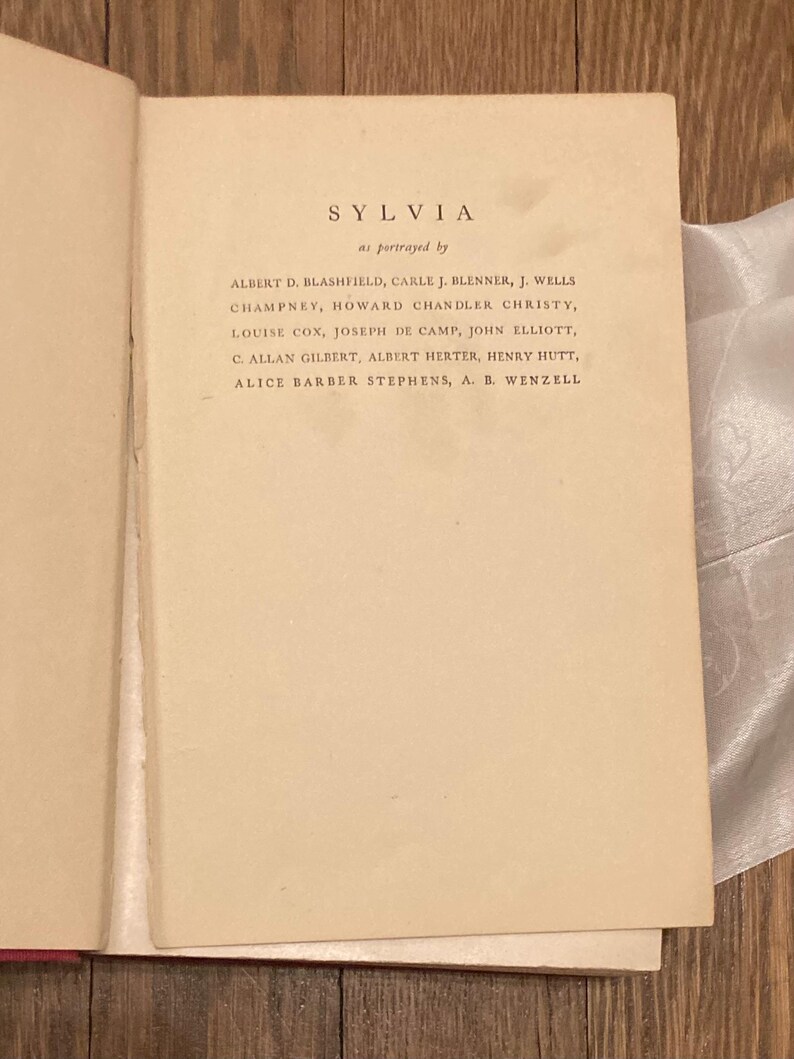 Puede incluir: P&aacute;gina de libro abierta con el t&iacute;tulo "SYLVIA" y una lista de nombres debajo, incluyendo Albert D. Blashfield y Howard Chandler Christy. El texto est&aacute; en una fuente serif en una p&aacute;gina color crema.