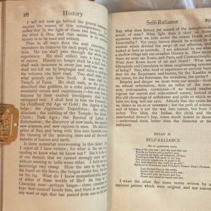 May include: Open book with text on pages 28 and 29, titled "History" and "Self-Reliance." A gold-colored bookmark with a decorative design rests on the left page. The pages are aged with a cream color.