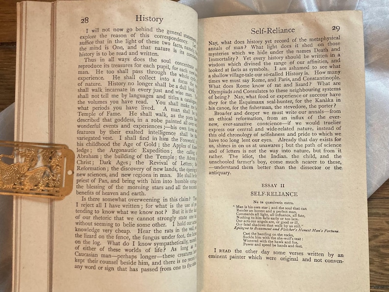 May include: Open book with text on pages 28 and 29, titled "History" and "Self-Reliance." A gold-colored bookmark with a decorative design rests on the left page. The pages are aged with a cream color.