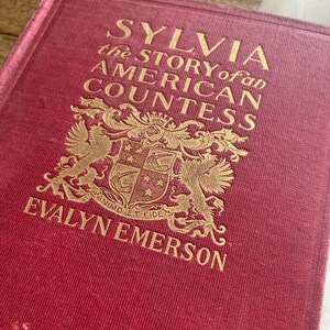 Puede incluir: Un libro de tapa dura rojo con letras doradas. El t&iacute;tulo es "SYLVIA the STORY of an AMERICAN COUNTESS" de Evalyn Emerson. La portada presenta un emblema detallado. El lomo del libro es visible, mostrando signos de desgaste.
