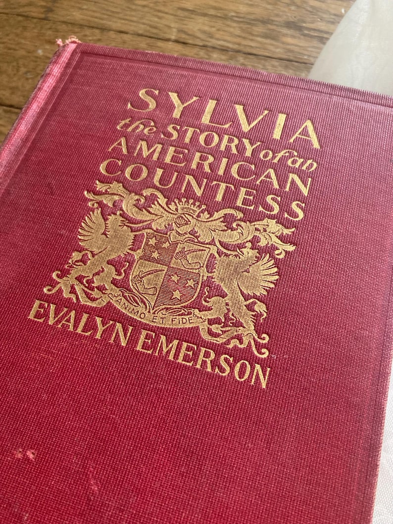 Puede incluir: Un libro de tapa dura rojo con letras doradas. El t&iacute;tulo es "SYLVIA the STORY of an AMERICAN COUNTESS" de Evalyn Emerson. La portada presenta un emblema detallado. El lomo del libro es visible, mostrando signos de desgaste.
