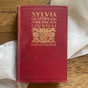 Puede incluir: Un libro antiguo titulado "Sylvia the Story of an American Countess" de Evalyn Emerson. El libro tiene una cubierta roja con letras doradas y un emblema decorativo. El libro est&aacute; sobre una tela blanca y una superficie de madera.
