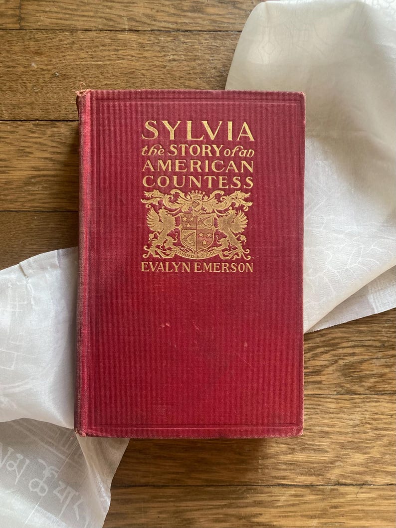 Puede incluir: Un libro antiguo titulado "Sylvia the Story of an American Countess" de Evalyn Emerson. El libro tiene una cubierta roja con letras doradas y un emblema decorativo. El libro est&aacute; sobre una tela blanca y una superficie de madera.