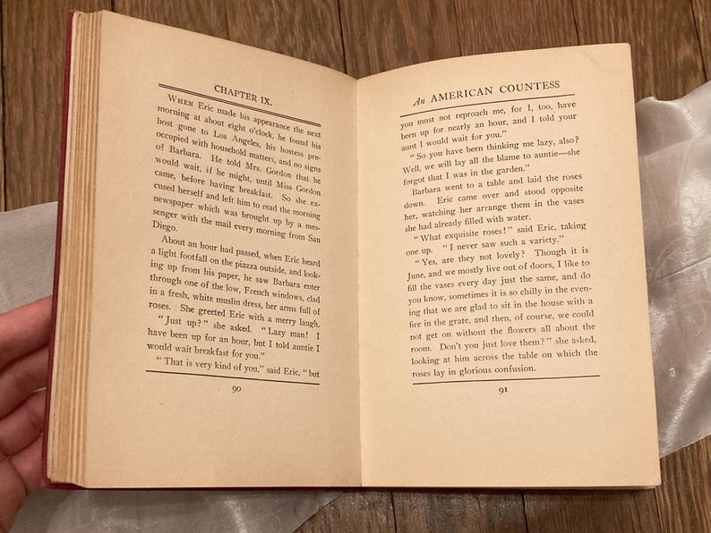 Puede incluir: Un libro abierto con p&aacute;ginas amarillentas y envejecidas. La p&aacute;gina izquierda muestra "CHAPTER IX." y la p&aacute;gina derecha dice "AN AMERICAN COUNTESS." El texto llena las p&aacute;ginas, con un lomo rojo visible.